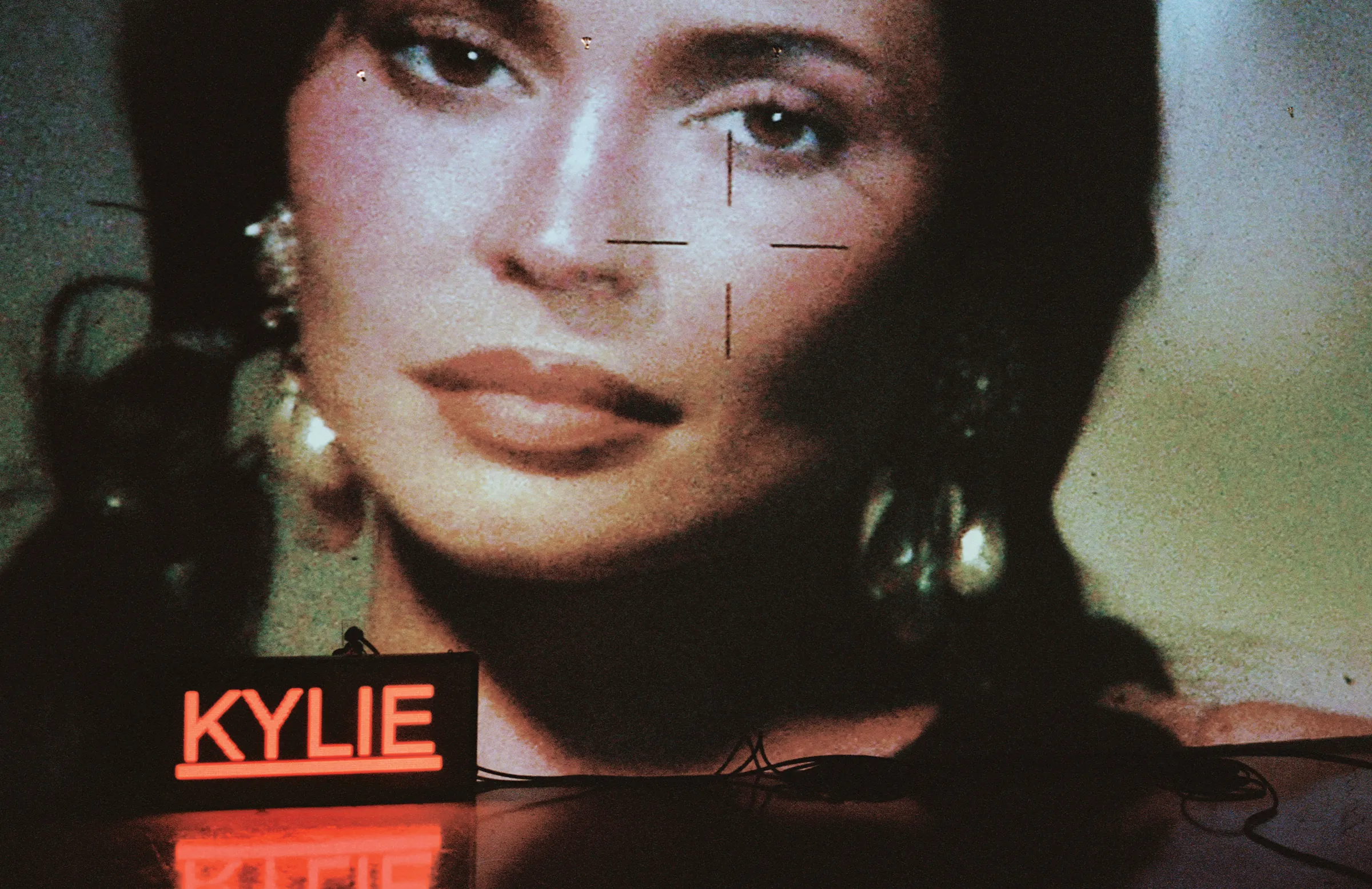 Close-up portrait of Kylie Jenner projected on a textured screen with a glowing neon Kylie sign below.