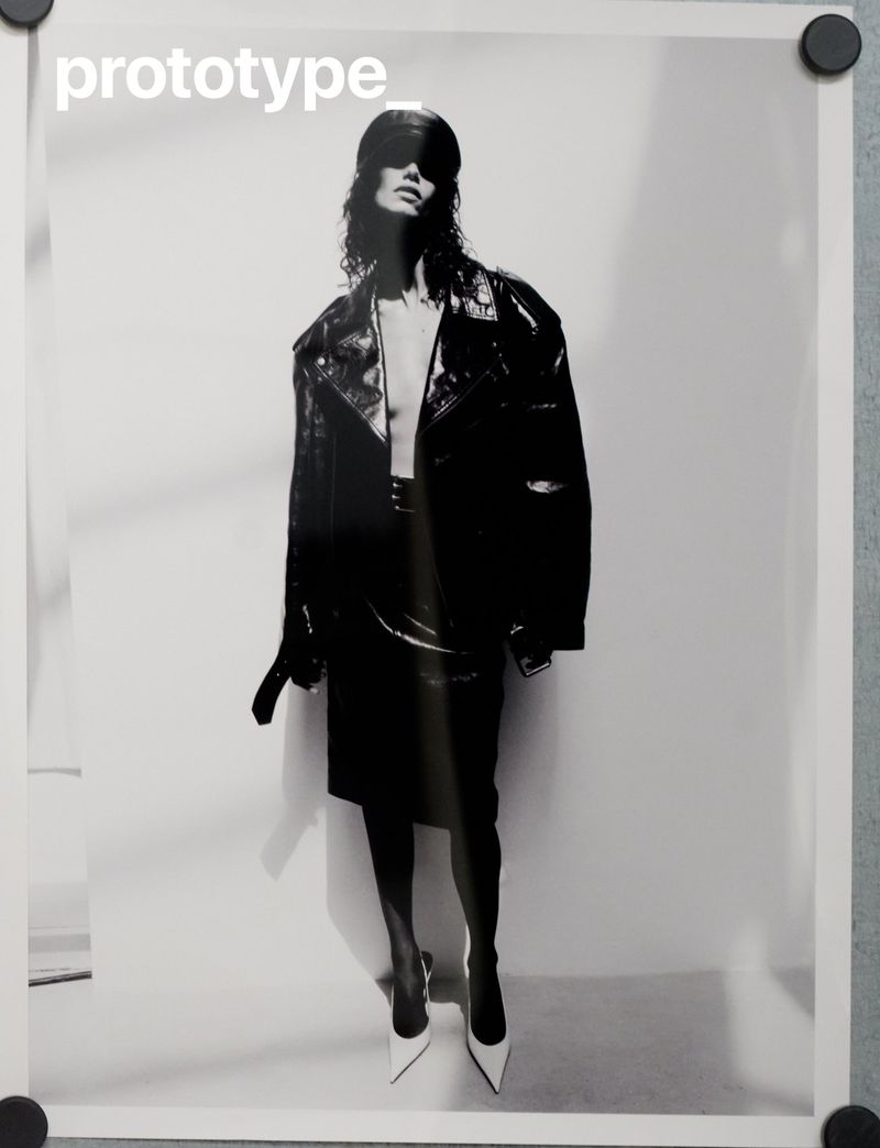Monochrome fashion portrait of a model wearing a glossy black leather jacket, skirt, cap, and white heels against a white background.