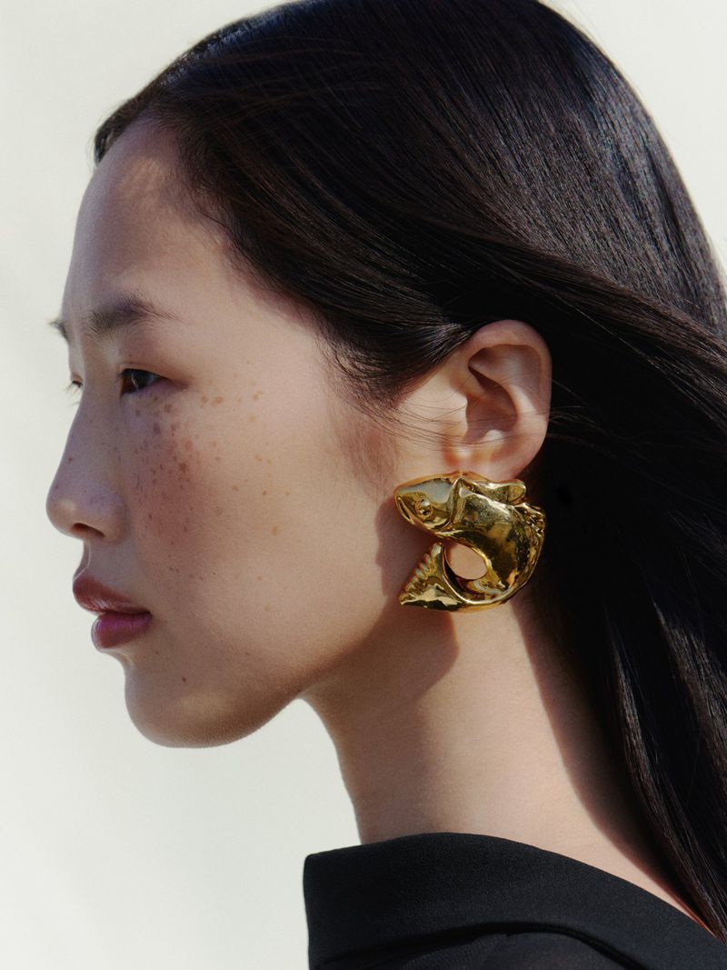 Profile portrait of a model with long dark hair wearing a large sculptural gold earring for Jacquemus FW26.