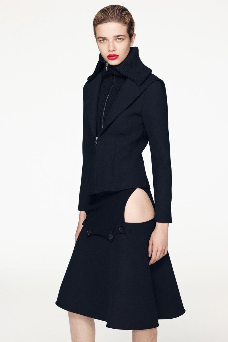 Model wearing a black zip-up structured jacket and cut-out skirt against a plain white studio backdrop.