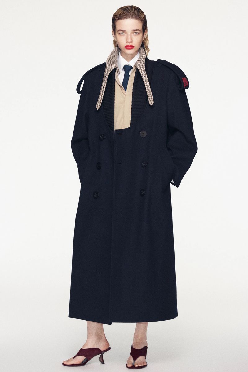 Model Lais Garcia wearing a long dark blue coat over a beige shirt and dark mules against a seamless white background.