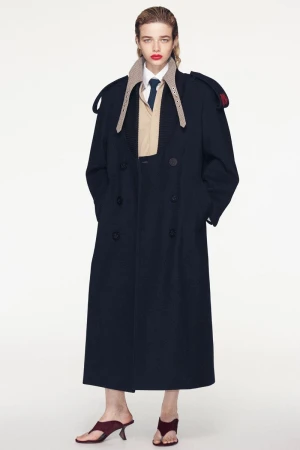Model Lais Garcia wearing a long dark blue coat over a beige shirt and dark mules against a seamless white background.