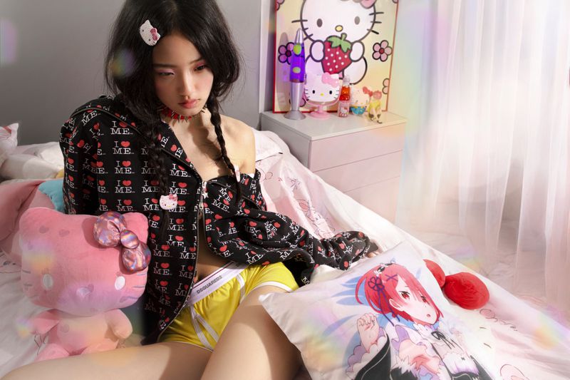 Young woman in bed wearing a patterned jacket, shorts, and Hello Kitty accessories, surrounded by plush toys and an anime pillow.