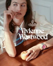 Model seated at a wooden table with an apple and a silver watch, wearing a white shirt and striped tie for Vivienne Westwood.