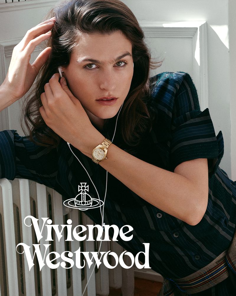 Model wearing a dark patterned shirt and gold wristwatch leaning against a white radiator in a light room