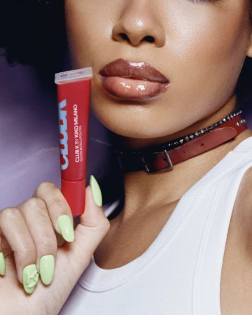 Close-up portrait of a woman holding a red Kiko Milano lipgloss tube, wearing a white top and brown glossy lips.