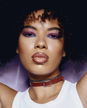 Close-up portrait of a woman with vibrant purple eyeshadow, glossy lips, dark curly hair, wearing a white top and a red buckle choker.