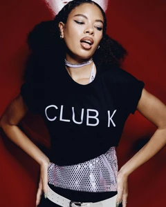 Woman in a black t-shirt with Club K text and a white belt posing against a rich red background