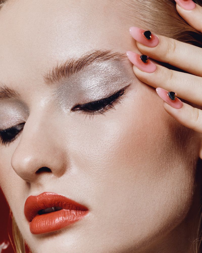 Close-up beauty portrait of a woman with metallic silver eyeshadow, red lipstick, and pink almond-shaped manicured nails.