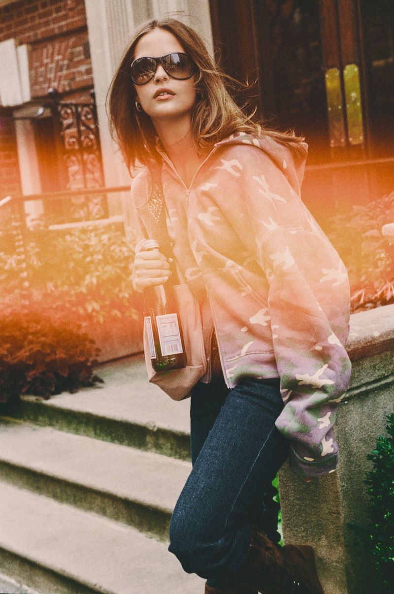 A young woman wearing sunglasses and a patterned zip-up hoodie standing on stone steps holding a bottle for Frankies Bikinis.