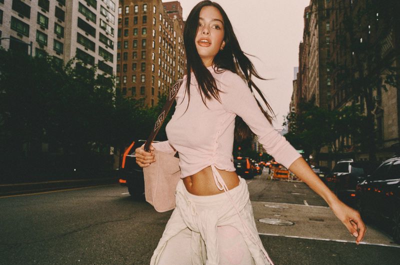 A long-haired woman in a light pink long-sleeve top and pale trousers walking down a city street with tall buildings in the background.