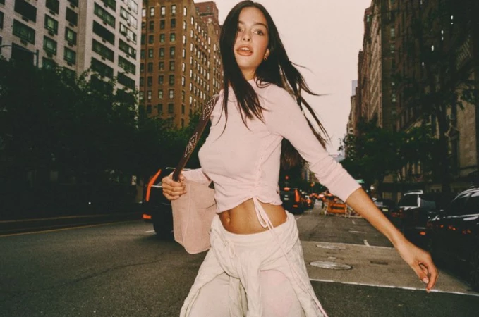 A long-haired woman in a light pink long-sleeve top and pale trousers walking down a city street with tall buildings in the background.