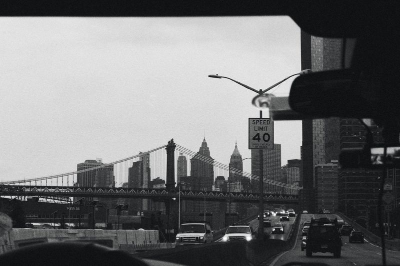 Black and white view looking through a car windshield at a city bridge and highway traffic with a speed limit sign