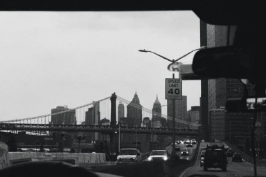 Black and white view looking through a car windshield at a city bridge and highway traffic with a speed limit sign