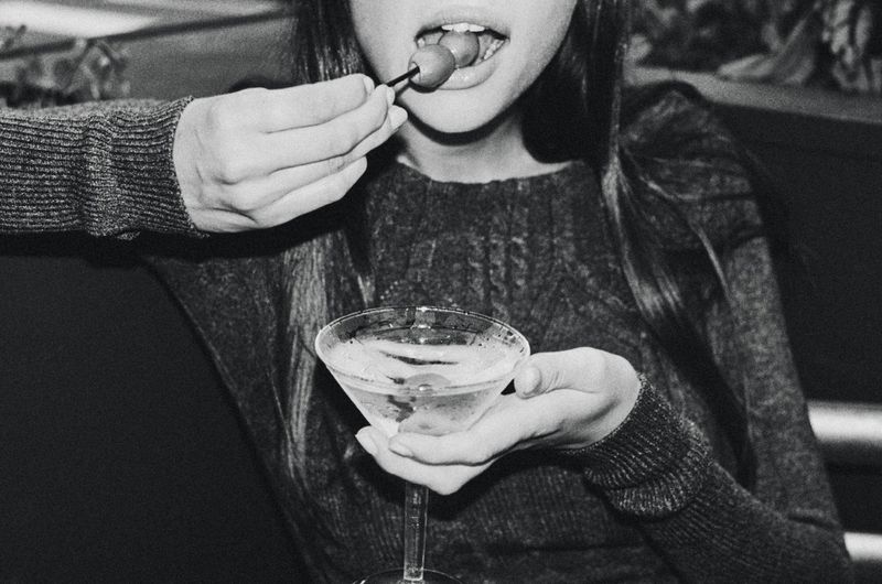 Monochrome close-up of a woman eating an olive from a toothpick while holding a martini glass.