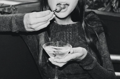 Monochrome close-up of a woman eating an olive from a toothpick while holding a martini glass.