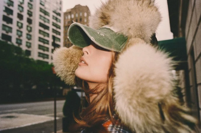 Close-up of a woman wearing a fluffy winter hat and green cap outdoors on a city street.