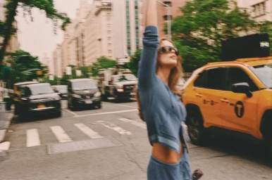 Woman in a blue knit zip-up hoodie raising her arm on a busy city street next to a yellow taxi