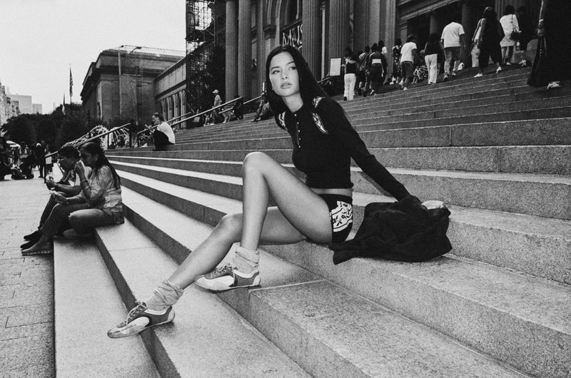 Model wearing dark long-sleeve knit top and shorts seated on wide stone steps outside a large public building