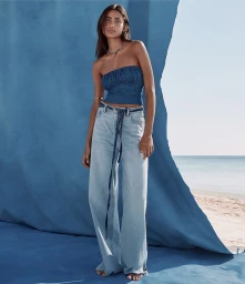 Model in a blue strapless top and light wash jeans posing before a blue textured backdrop with a sandy beach visible in the background.