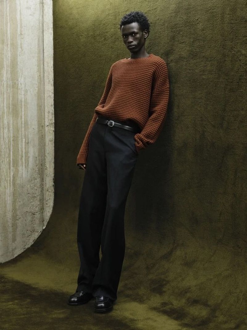 Black man wearing a rust-orange sweater and black trousers leaning against an olive textured studio backdrop for Fursac F/W 2026 campaign