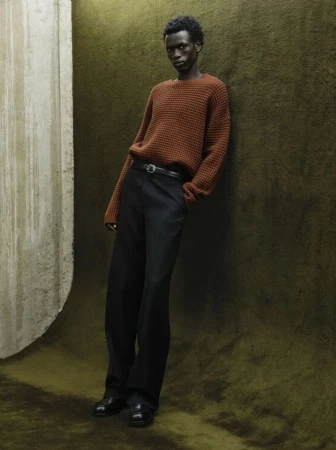 Black man wearing a rust-orange sweater and black trousers leaning against an olive textured studio backdrop for Fursac F/W 2026 campaign