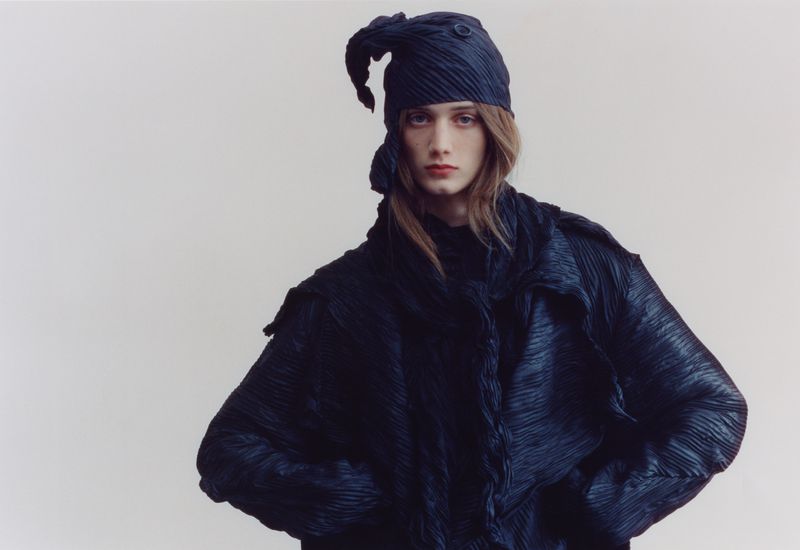 Model in pleated dark jacket and matching headwrap against a plain white background for The Blend Magazine