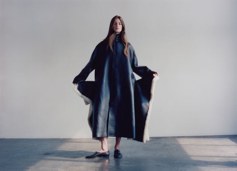 Model standing against a neutral wall wearing a long dark coat with white lining and black shoes