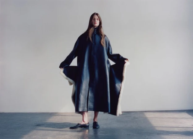 Model standing against a neutral wall wearing a long dark coat with white lining and black shoes