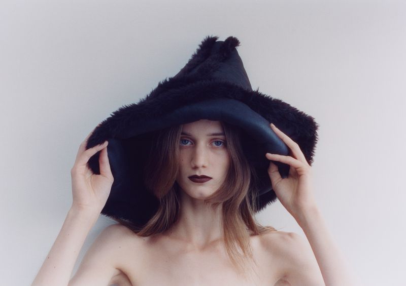 Model wearing a large black furry hat holding the brim against a plain white background for The Blend Magazine