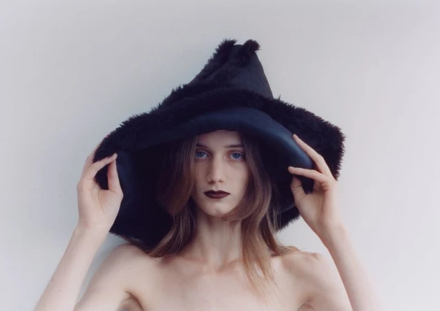 Model wearing a large black furry hat holding the brim against a plain white background for The Blend Magazine