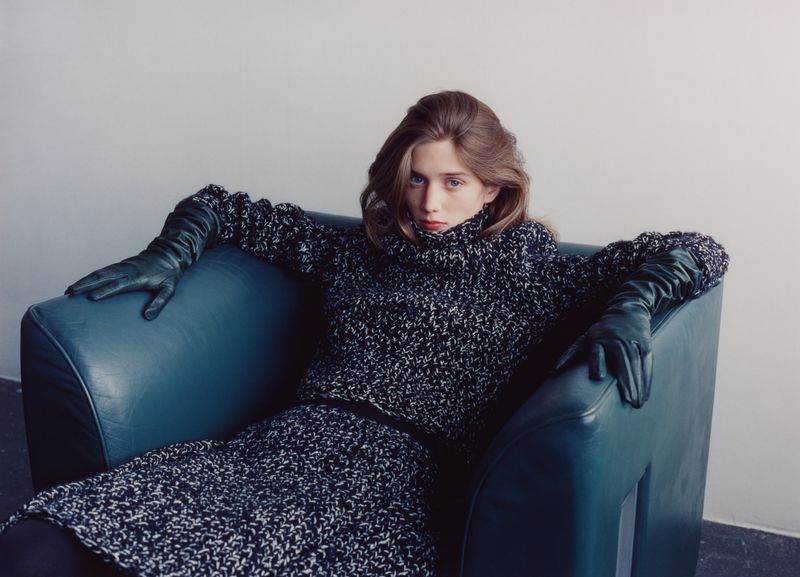 Model seated in a dark armchair wearing a textured black and white turtleneck sweater and dark green leather gloves.