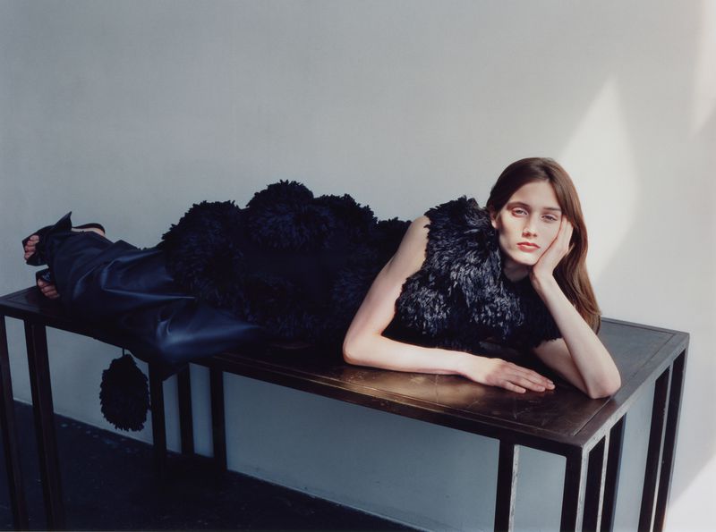 Model lying on a dark table wearing a black textured top against a plain wall