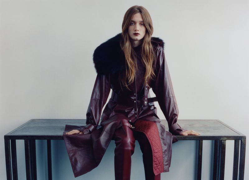 Model seated on a metal table wearing a burgundy leather coat with a dark fur collar