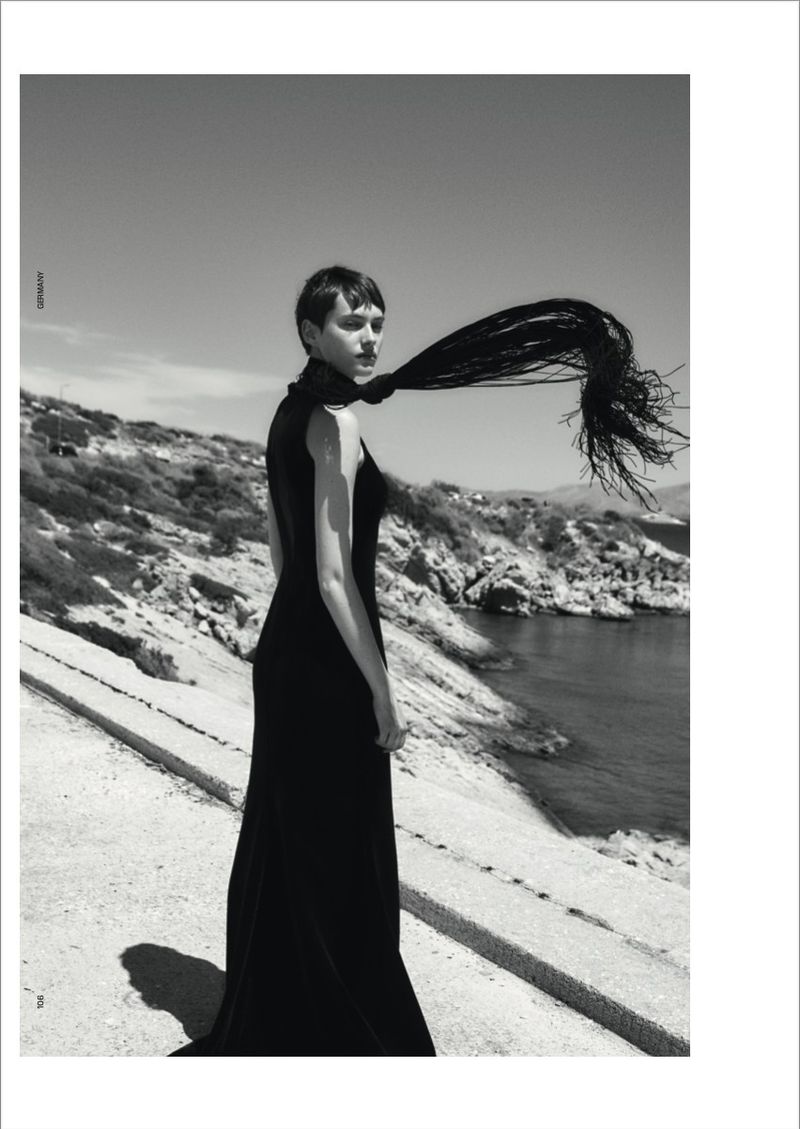 Model Greta Hofer in a black gown with wind-swept hair standing outdoors by the sea for T Magazine Germany