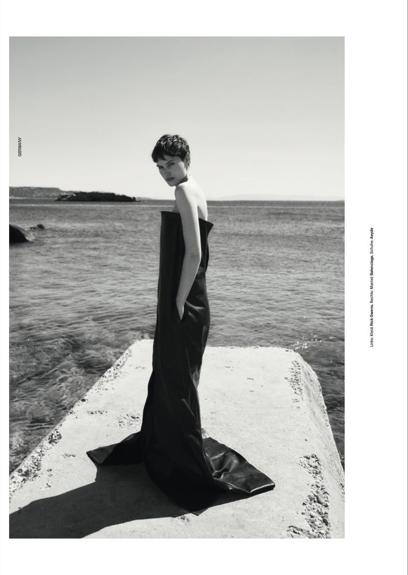 Model with short dark hair wearing a long black strapless dress standing on a concrete pier overlooking the sea.