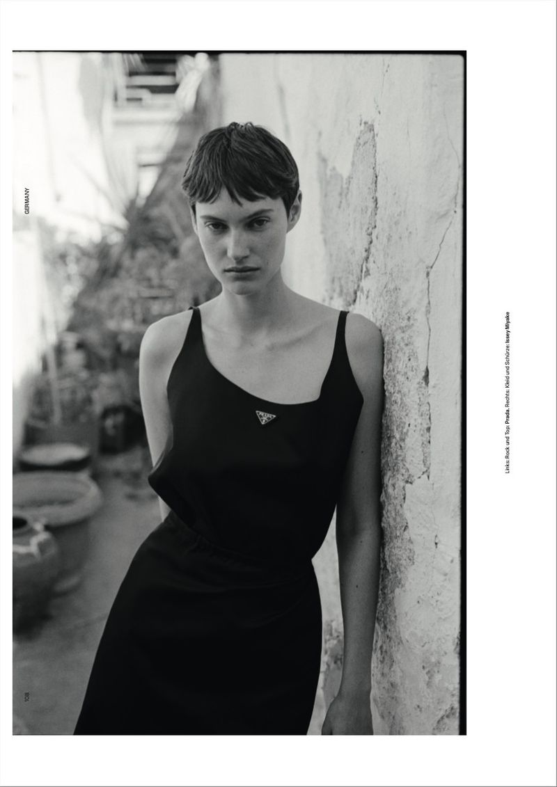 Monochrome portrait of a female model in a black dress leaning against a textured stone wall in an outdoor courtyard.