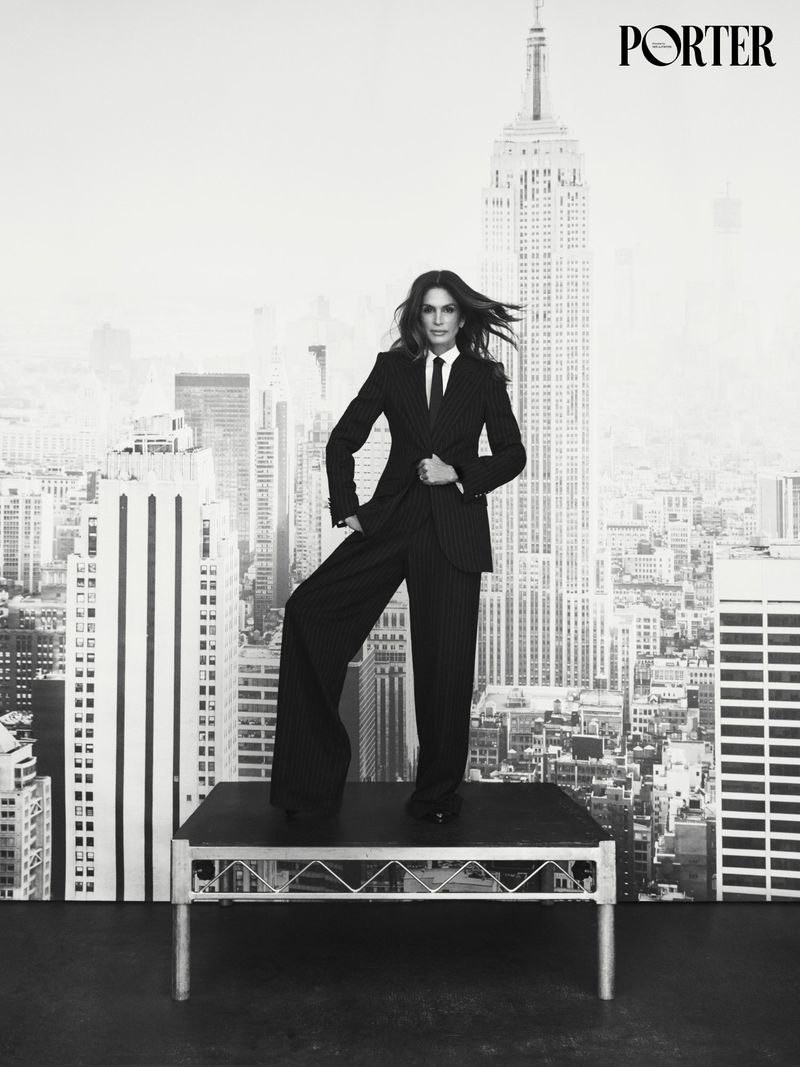 Cindy Crawford wearing a pinstripe suit standing on a raised platform against a city skyline backdrop.