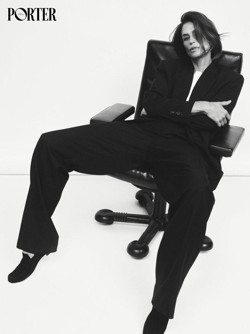 Cindy Crawford sitting in a black office chair wearing a black suit jacket and trousers against a white backdrop.