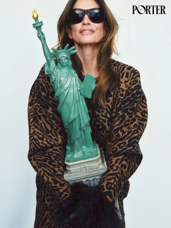Cindy Crawford wearing sunglasses and an animal print coat holding a Statue of Liberty prop against white seamless background