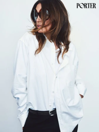 Cindy Crawford wearing oversized sunglasses and a white button-down shirt against a clean white backdrop