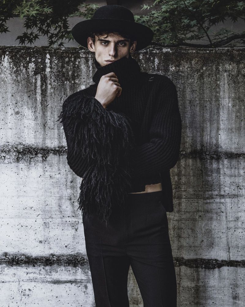 A male model wearing a black hat, black turtleneck, and trousers against a textured concrete wall.
