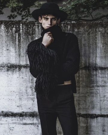A male model wearing a black hat, black turtleneck, and trousers against a textured concrete wall.