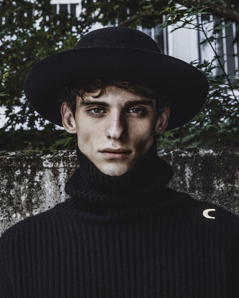 Close-up portrait of a man wearing a black wide-brimmed hat and black ribbed turtleneck sweater against an outdoor wall.
