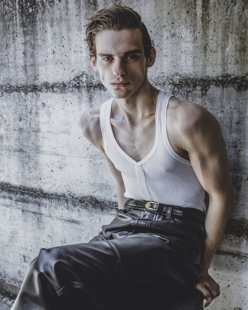 A young man wearing a white tank top and dark leather pants seated against a weathered concrete wall.