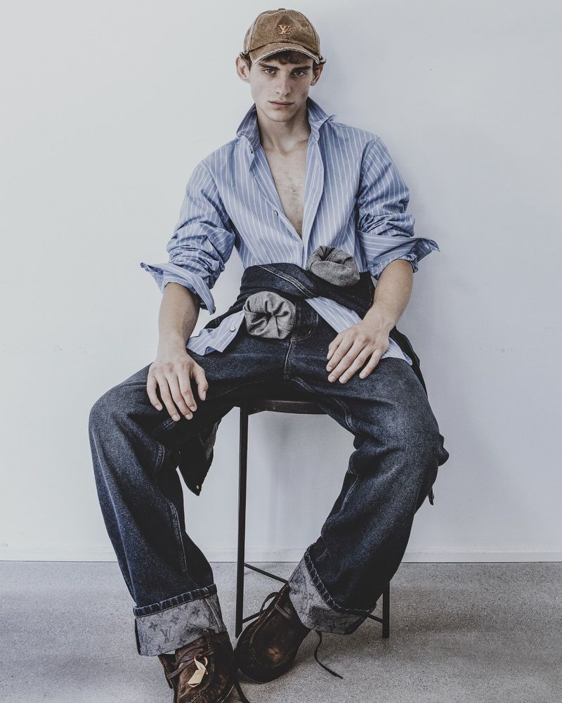 A young male model wearing a striped shirt and dark denim jeans seated on a stool against a white background.