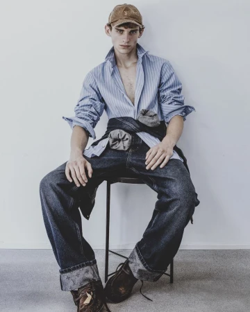 A young male model wearing a striped shirt and dark denim jeans seated on a stool against a white background.