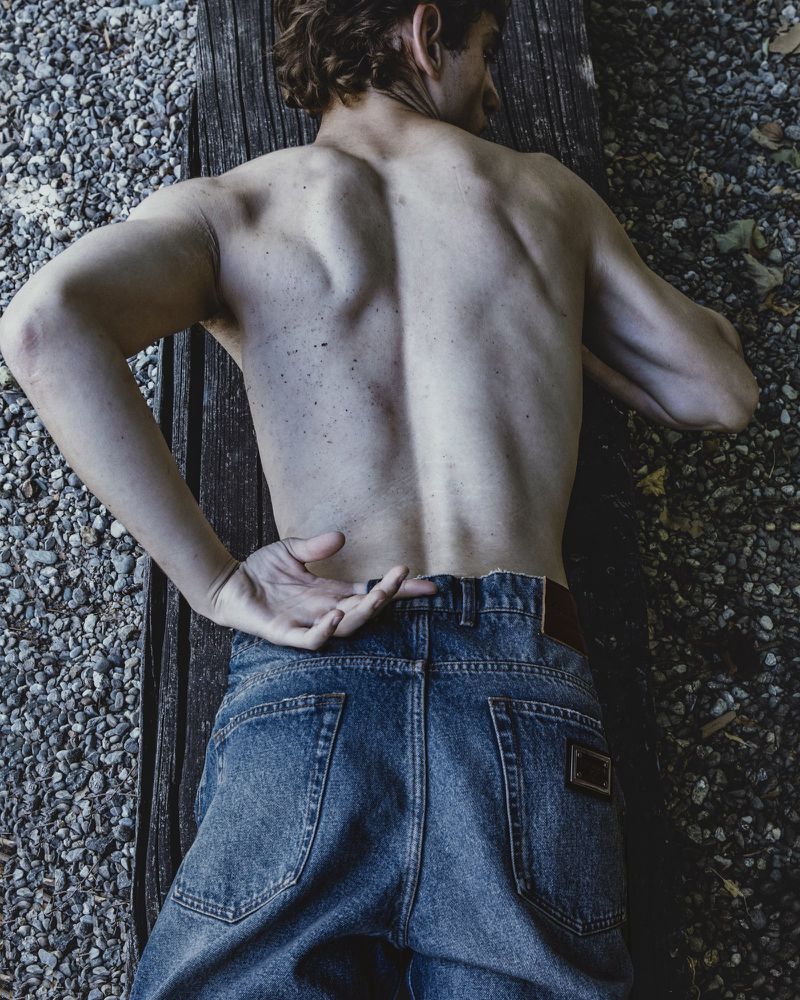 Shirtless man lying face down on a weathered wooden bench wearing blue denim jeans over a gravel ground