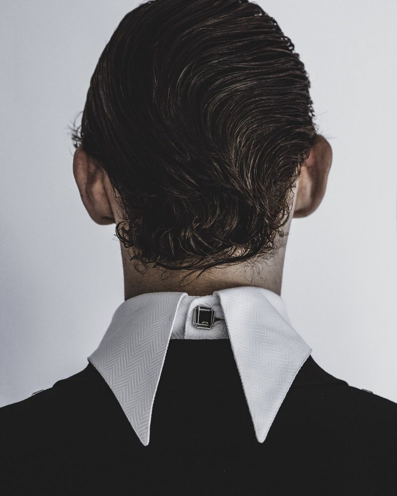 Rear view of a person wearing a white stiff-collared shirt and black jacket against a plain grey backdrop.