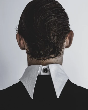 Rear view of a person wearing a white stiff-collared shirt and black jacket against a plain grey backdrop.